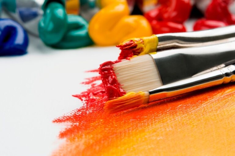 Close-up of paintbrushes loaded with red acrylic paint resting on a canvas streaked with orange, with blurred paint tubes in the background.