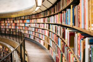 Curved multi-tiered library bookshelves lined with colorful books, receding around a balcony railing beneath a curved ceiling.