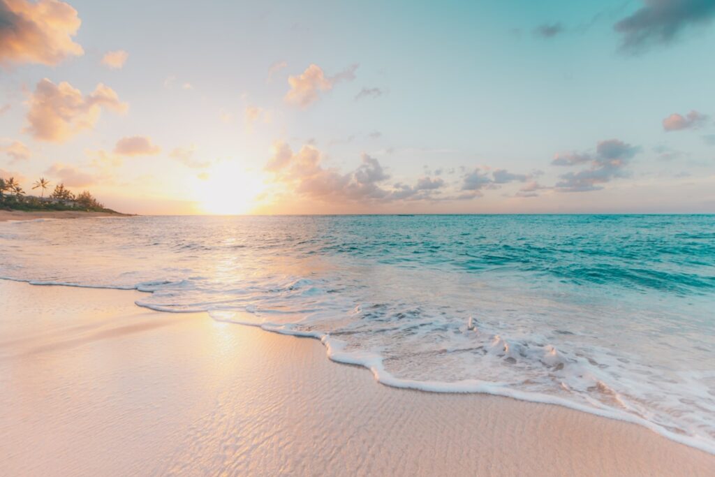 Tropical beach at sunset with turquoise waves lapping onto pale sand and distant palm trees on the left horizon.