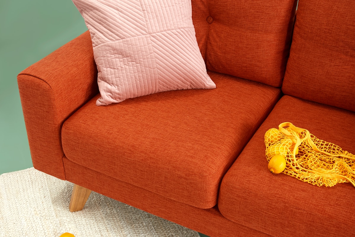 Close-up of a terracotta-orange upholstered sofa with a quilted pale pink cushion and a yellow mesh bag of lemons, against a sage green wall.