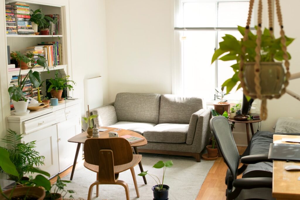 Small sunlit living room corner with a gray sofa, white bookshelf full of books, mid-century wood coffee table, and many leafy houseplants.