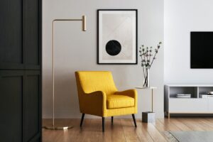 Minimalist living room with a mustard-yellow armchair, brass arc floor lamp, and a framed abstract print featuring a black circle, on a pale wood floor.