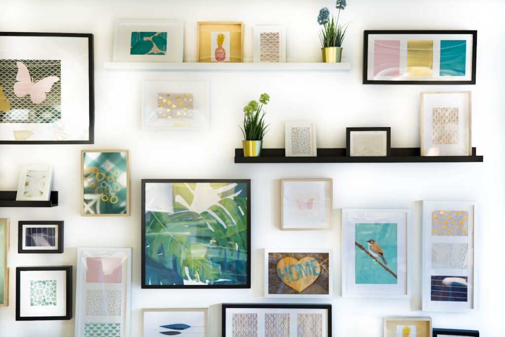 Dense gallery wall of many small framed prints and pastel artworks on picture ledges, with a large framed tropical-leaf print at the center, on a white wall.