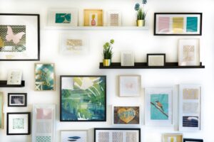 Dense gallery wall of many small framed prints and pastel artworks on picture ledges, with a large framed tropical-leaf print at the center, on a white wall.