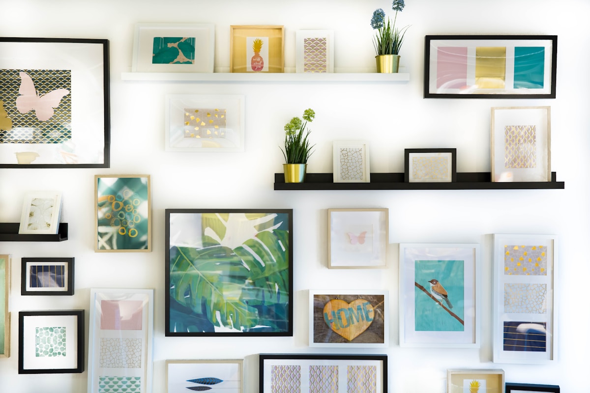 Dense gallery wall of many small framed prints and pastel artworks on picture ledges, with a large framed tropical-leaf print at the center, on a white wall.