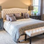 Elegant bedroom with a tufted cream upholstered headboard, silver sequined throw, gray and purple pillows, and mirrored studded nightstands with tall lamps.