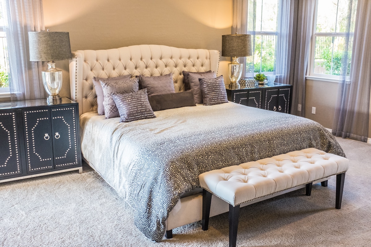 Elegant bedroom with a tufted cream upholstered headboard, silver sequined throw, gray and purple pillows, and mirrored studded nightstands with tall lamps.
