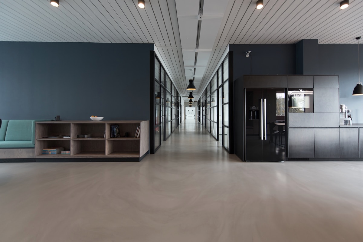 Empty modern open-plan office corridor with polished concrete floor, a glass-walled meeting room on the right, and a long wooden credenza on the left.