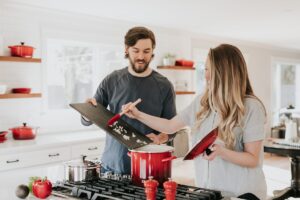 A man and a woman cooking together at a gas range in a bright white kitchen, tending to a red enamel pot with a wooden spoon.