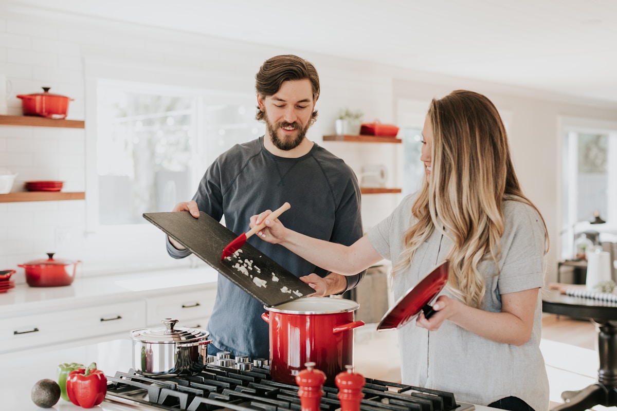 A man and a woman cooking together at a gas range in a bright white kitchen, tending to a red enamel pot with a wooden spoon.