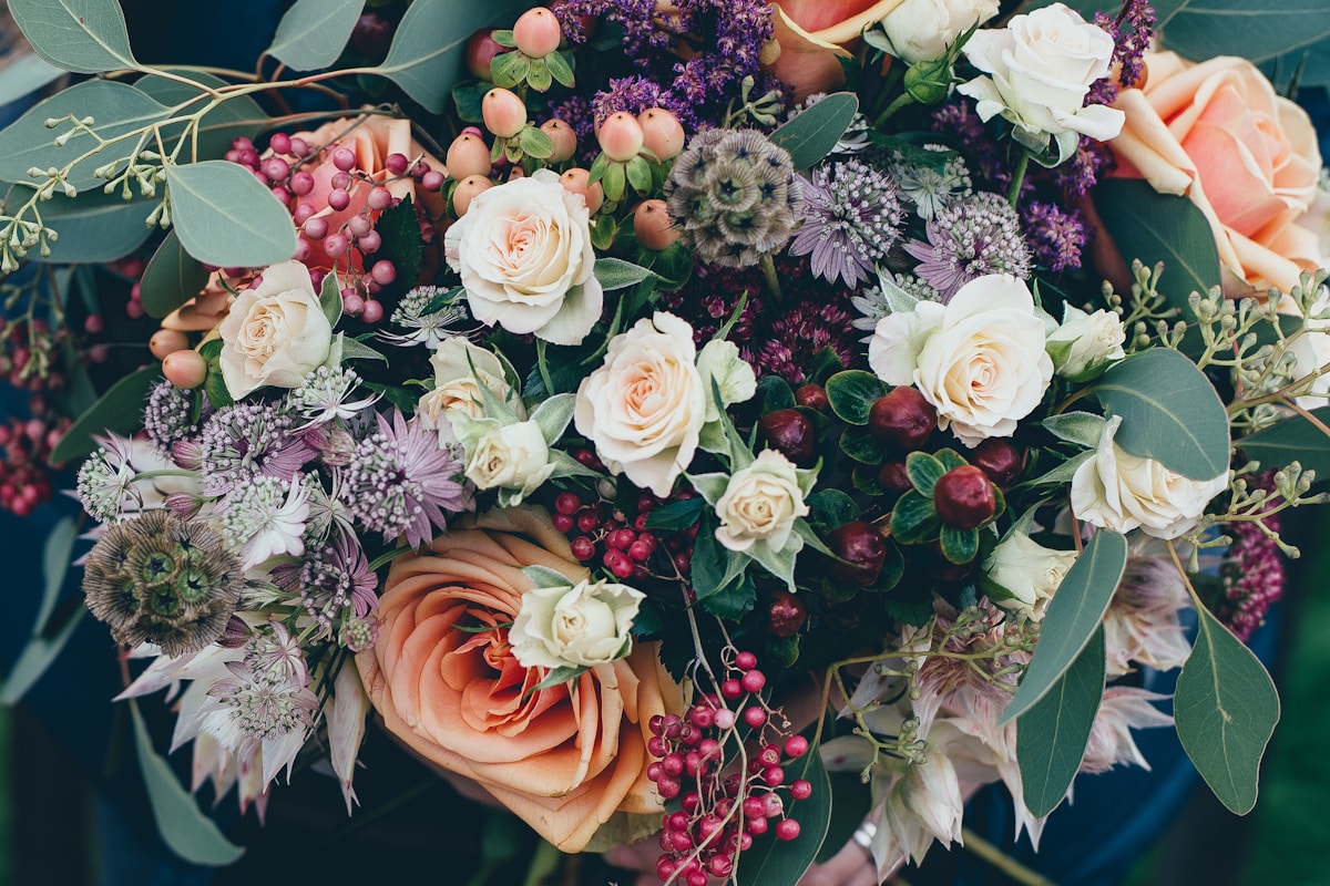 Close-up of a full floral bouquet with peach and cream garden roses, deep purple berries, pink hypericum, and eucalyptus foliage.
